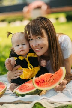 Happy mother and baby girl eating fresh red watermelon slice on green grass outdoors picnic, family summer concept.