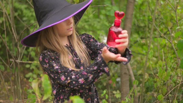 girl in black witch hat holding red glass potion bottle with heart stopper, little sorceress preparing love spell in magic forest, first love, cute childhood fantasy story, pagan witchcraft ritual