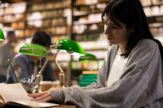 Focused young woman reading from open book at library study table with laptop and green lamps, warm lighting and bookshelves in university library for academic research and self-study.
