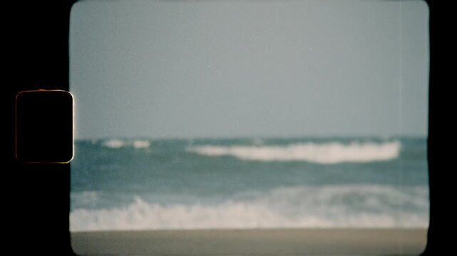 Young man walking along the sandy beach shoreline near the ocean waves, captured in a vintage Super 8 film style with sprocket holes visible on the side