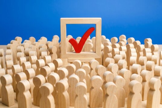 A crowd of wooden people figures looking at a red checkmark in a wooden box for voting elections.