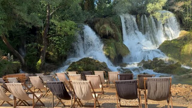 Several empty wooden lounge chairs are neatly arranged in rows near a natural waterfall cascade, suggesting a resort or park setting