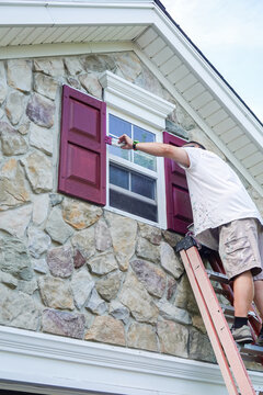 Handyman on a ladder painting the shutters next to a small window over a garage