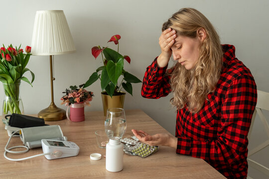 Confused sick woman choosing medication from many pills while touching her forehead at home. Concept of self-treatment uncertainty, headache, polypharmacy and risk of incorrect drug use.
