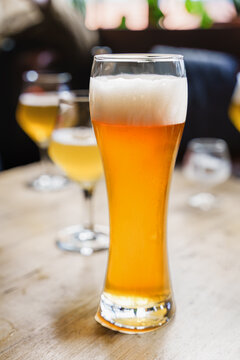 Pint glass of golden beer with foamy head on wooden table in relaxed pub setting, with blurred glasses behind it, creating cheerful social drink presentation and warm bar atmosphere