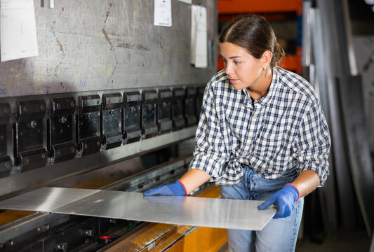 Young woman worker bends sheet of metal under press in metallurgical workshop