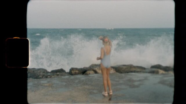 Girl in a swimsuit stands on a rocky shore looking at ocean waves, captured in slow motion on vintage Super 8 film