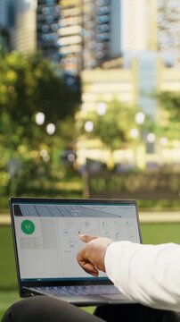 Portrait format: Businessman checks solar energy metrics and system data on a laptop outdoors in a city park, using high-tech sustainability data for a photovoltaics project.