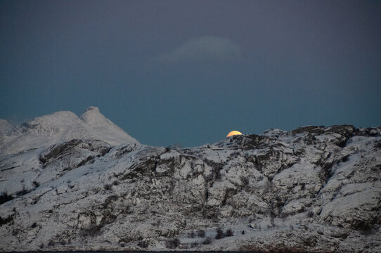 A near full moon is slowly creeping behind a mountain along the norwegian coast on an early winter morning near the town of Bodo.
