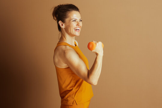 A smiling, fit middle-aged woman in orange activewear, hair in bun, holds dumbbell in bicep curl pose. Her joyful expression against light brown background conveys energy, confidence and well-being