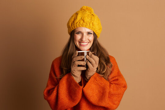 An adult woman with long hair, wearing yellow knitted pom-pom beanie and orange sweater, smiles directly at camera while holding brown mug. The cozy scene conveys cheerful autumn or winter atmosphere