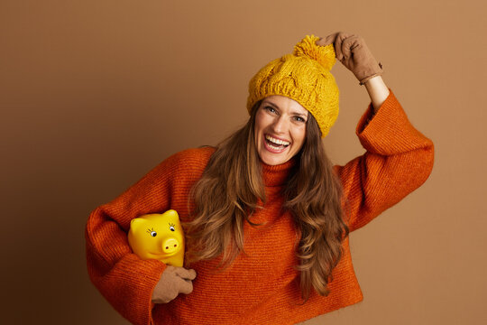 A joyful woman in orange sweater and yellow beanie laughs heartily while holding yellow piggy bank in her gloved hands. This image symbolizes financial planning and saving during cozy autumn season