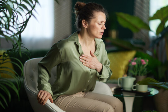 A woman in a green satin blouse clutches her chest with an expression of distress, illustrating symptoms of anxiety, heart pain, or a panic attack at home.