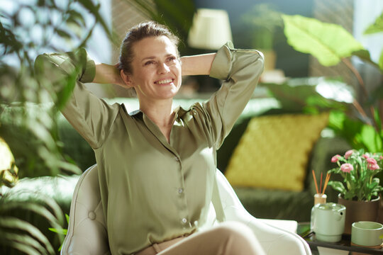 A woman finds balance and peace at home, smiling as she relaxes in a bright, plant-filled room with a teapot nearby.