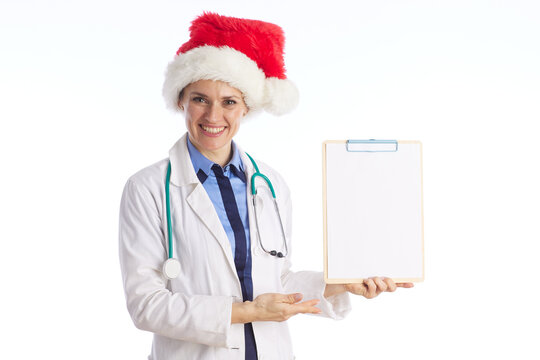 A smiling female doctor in a white lab coat and Santa hat holds a blank clipboard, blending professional medical care with festive holiday cheer on a white background.