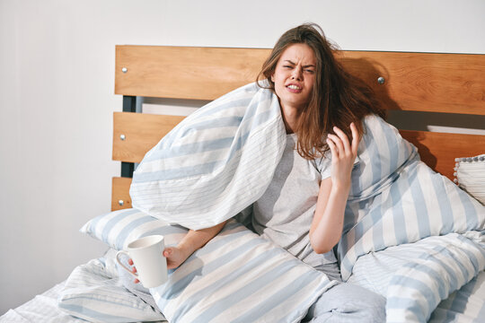 Sick young woman waking up in bed under a striped blanket, holding a mug and looking tired and unwell in the morning at home.
