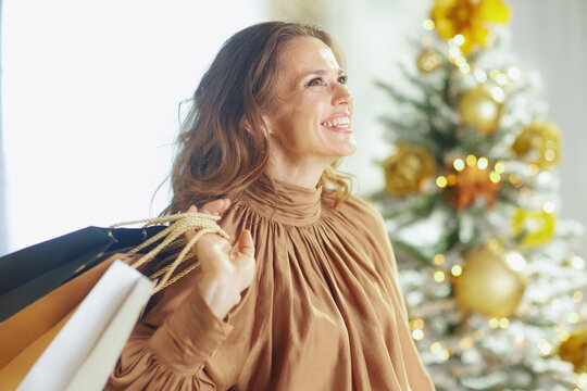 Radiant woman in gold satin turtleneck beams, carrying shopping bags. Softly blurred Christmas tree with gold ornaments and fairy lights creates bright, festive holiday atmosphere.