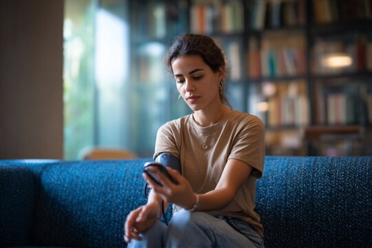 Young woman checks her blood pressure on a couch in a bright room with bookshelves
