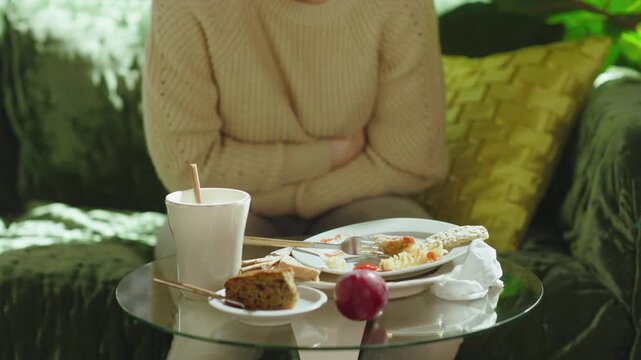 A woman sits on a green sofa clutching her stomach and hunching forward in distress, depicting acute abdominal pain or food poisoning after a meal.
