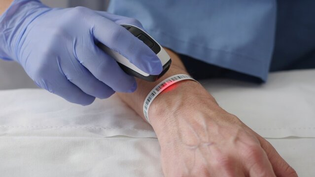 Barcode scanner in a gloved hand reading a patient wristband on an arm inside a clinic