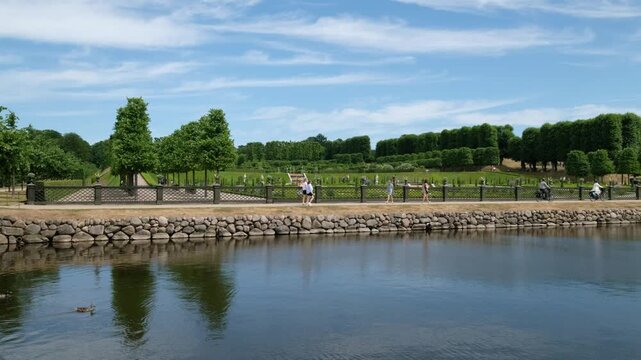 A picturesque view of Frederiksborg Castle grounds in Denmark. See people walk and bike along a lakeside path. Calm and serene
