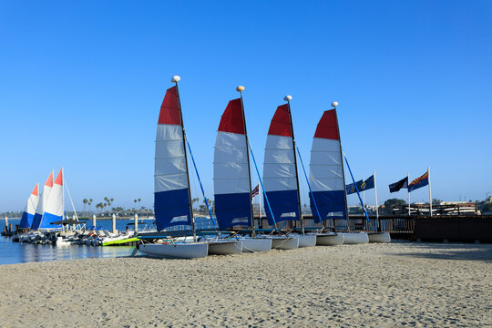 Row of small catamarans with red white and blue sails parked on a Mission Bay sandy beach under a clear blue sky, San Diego, CA