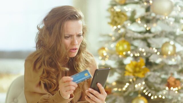 A middle-aged woman with wavy brown hair sits with distressed expression, holding credit card and smartphone. A decorated Christmas tree in background highlights financial stress during holiday season