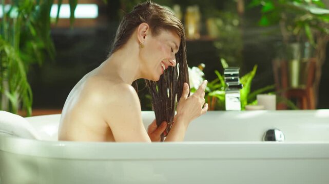 A woman with brown hair smiles, examining wet hair while relaxing in a white bathtub. Surrounded by green plants & white flowers, it offers a tranquil, spa-like setting for her beauty routine.