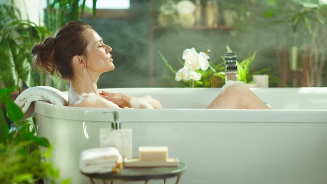 A woman indulges in a peaceful, steamy bubble bath within a lush, plant-adorned bathroom, embracing a moment of self-care and serenity in her home spa.