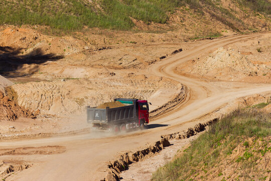Heavy red tipper truck transporting sand along a winding dirt road in a sunny open-pit quarry during early autumn: Russia - September 9, 2026
