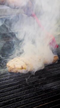 A piece of pork shoulder cooks on a black grill while a hand brushes oil over the meat. As the oil reaches the hot surface, thick smoke rises and intense flames suddenly flare around the pork, creatin