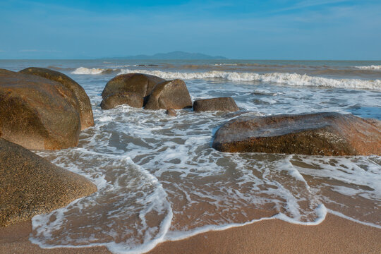 Waves crash against large dark rocks on coastal beach under clear blue sky