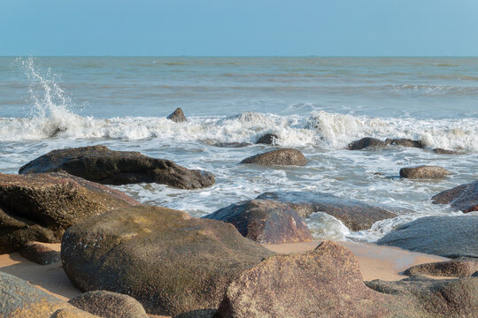 Waves crash against large dark rocks on coastal beach under clear blue sky