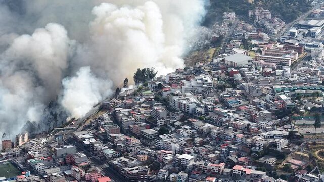 Aerial view of a large fire and thick smoke rising above Quito urban neighborhoods, roads, sports fields and a wooded ravine in Ecuador.