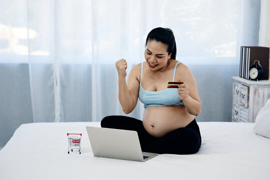 Happy pregnant adult woman shopping online with laptop, credit card and cart on bed, celebrating successful payment, ecommerce purchase and convenient home retail