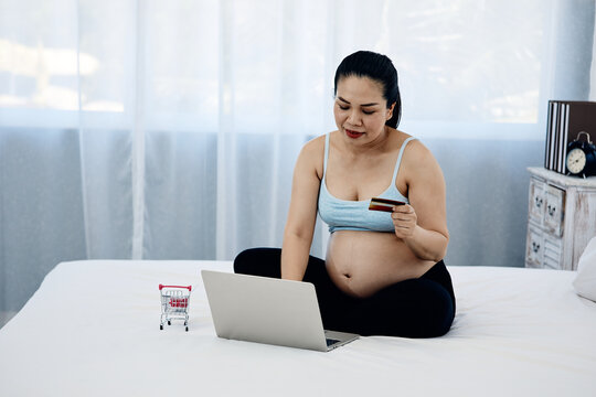 Pregnant adult woman shopping online with laptop, credit card and miniature cart on bed, showing maternity lifestyle, ecommerce, payment and home convenience