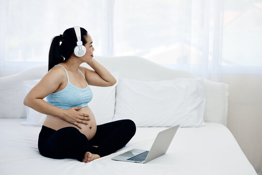Pregnant woman wearing headphones while touching belly beside laptop on bed, enjoying music, maternity relaxation, wellness, bonding and peaceful home lifestyle