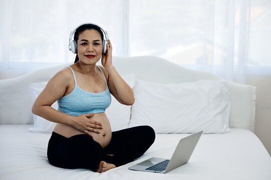 Pregnant woman wearing headphones beside laptop while touching belly and smiling at camera on bed, enjoying music, maternity wellness, relaxation, bonding and peaceful home lifestyle