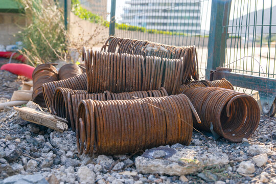 A group of large rusty coiled steel springs sit stacked on gravel behind a wire fence at a scrapyard, a concept of industrial waste and metal recycling.