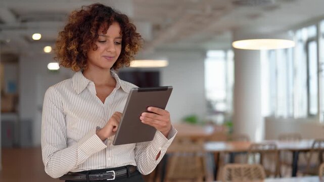Young hispanic business woman using digital tablet in modern coworking office, scrolling touchscreen. Smiling latin manager browsing company report online, checking market data on mobile device