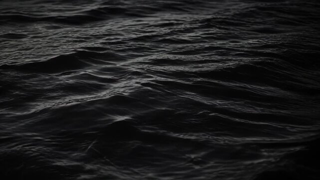Dark ocean surface with rolling sea wave movement, rippling water texture, subtle highlights, and dramatic low light creating moody abstract background for nature design and atmospheric visuals