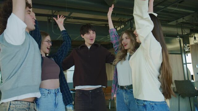 A group of people stands in a circle indoors, stacking their hands together in the center as a gesture of unity and teamwork.