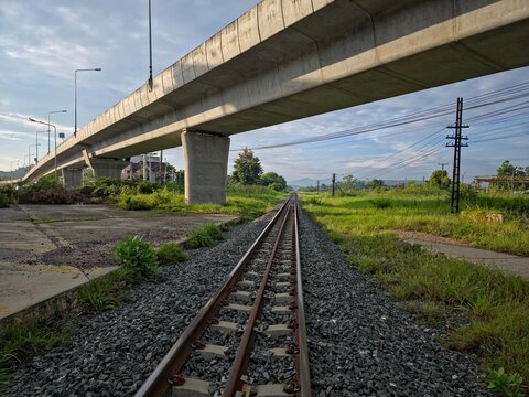 Railway track extending beneath a massive elevated concrete viaduct through a green countryside landscape, featuring utility poles, distant mountains, and strong leading lines toward the horizon in so