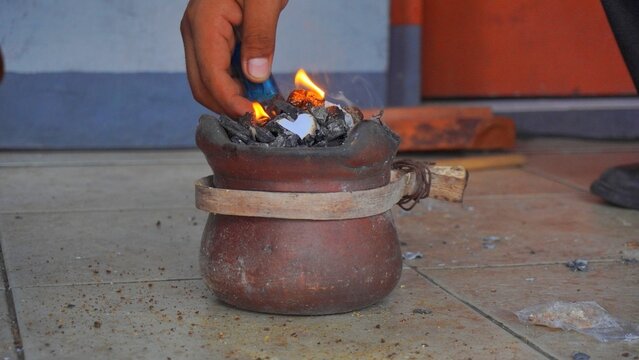 Close-up of a person's hand using a lighter to ignite charcoal in a traditional terracotta stove on a tiled floor