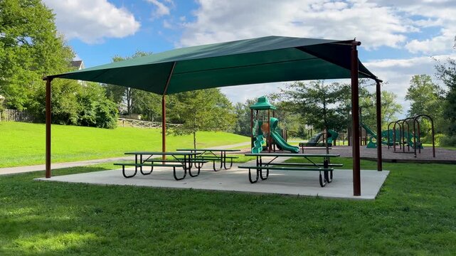 Picnic shelter with tables on a concrete pad next to a playground in bright daylight. Horizontal video featuring vibrant park aesthetics, green shade sail, and peaceful summer environment.