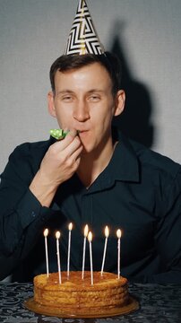 Cheerful caucasian man wearing a party hat celebrating his birthday alone, clapping hands, making a wish, and blowing a noisemaker while sitting in front of a birthday cake with candles