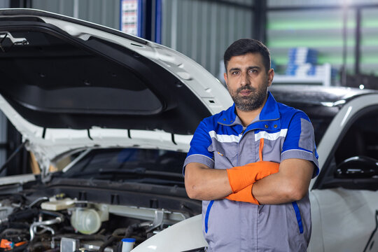 Man professional automotive technician standing with crossed arms beside car with open hood in workshop, representing vehicle maintenance, repair service, skilled labor and mechanic expertise.