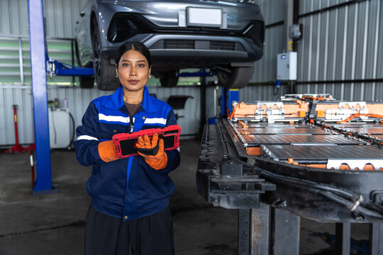 Female electric vehicle technician holding digital tablet beside EV battery pack in automotive workshop, representing battery service, skilled workforce, engineering and clean mobility technology.