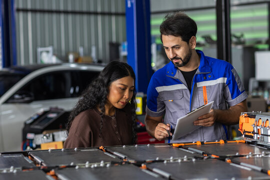 Man Electric vehicle technician explaining EV battery pack to female customer in an automotive workshop, showing battery service, technical consultation, maintenance and electric car technology.