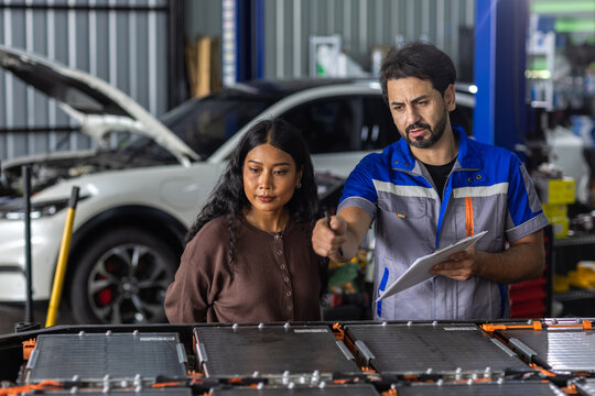 Man Electric vehicle technician explaining EV battery pack to female customer in an automotive workshop, showing battery service, technical consultation, maintenance and electric car technology.